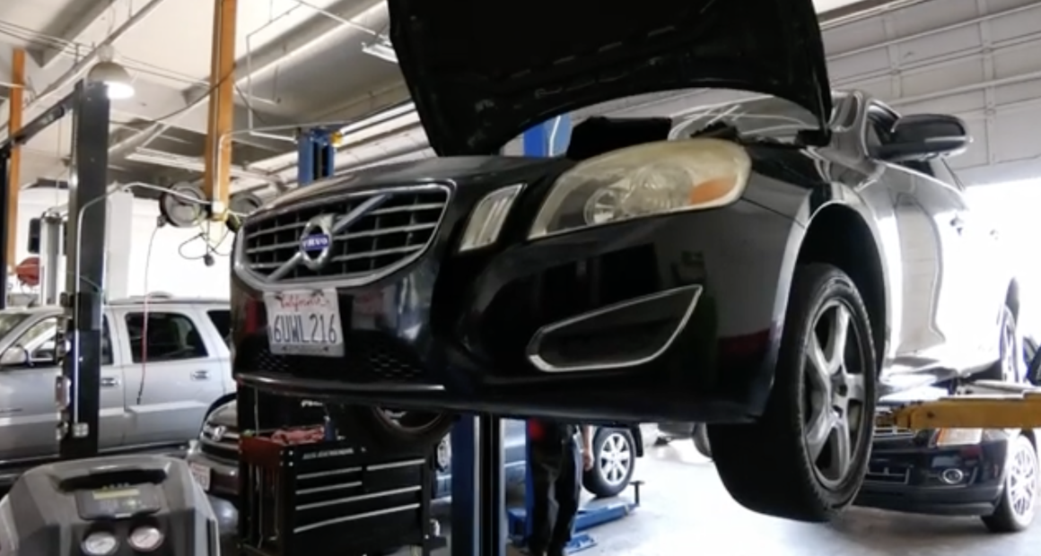 A black Volvo sedan with its hood open is lifted on a hydraulic car lift inside an auto repair shop. The vehicle has a California license plate and is surrounded by other cars and equipment, indicating a busy maintenance environment.